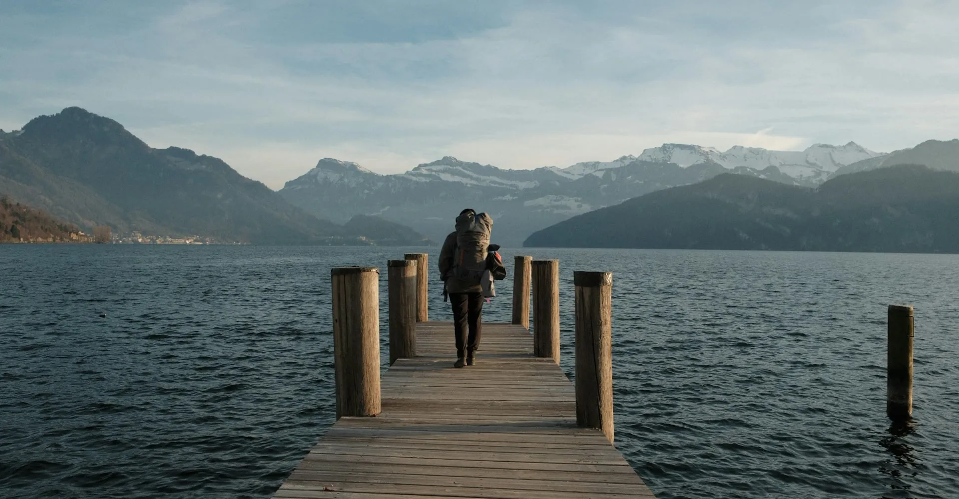 A person standing on a wooden jetty beside a lake and mountains